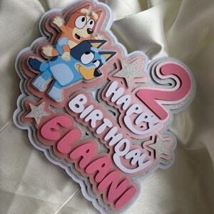 May include: A layered cake topper featuring the cartoon characters Bluey and Bingo, the number 2 in pink, and the text Happy Birthday Elaani in white and pink. The design includes silver glitter accents and star shapes on a white base.