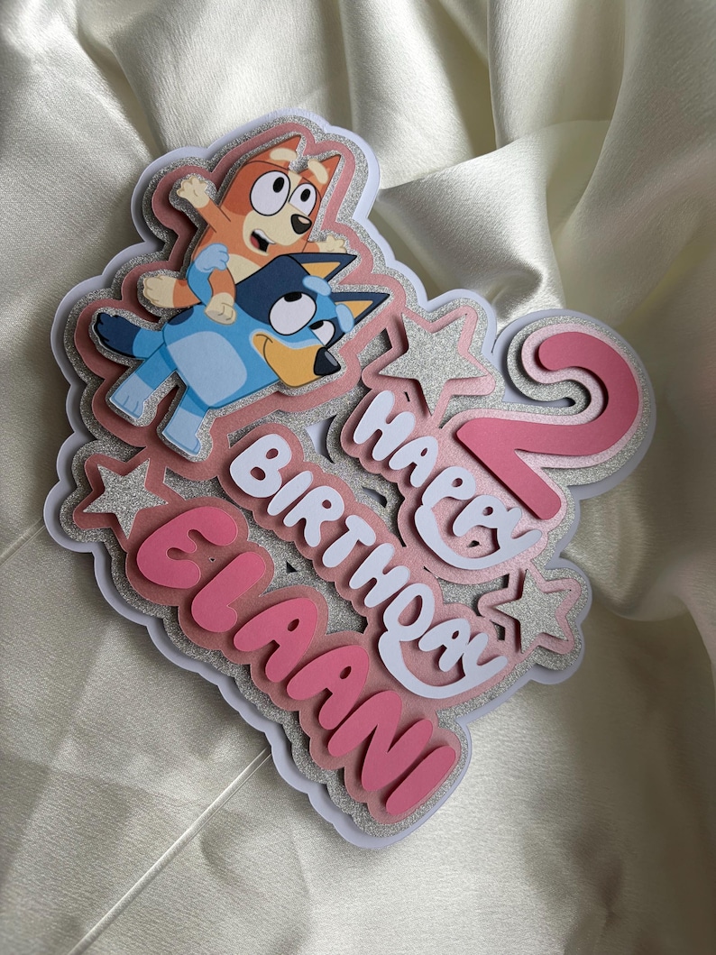 May include: A layered cake topper featuring the cartoon characters Bluey and Bingo, the number 2 in pink, and the text Happy Birthday Elaani in white and pink. The design includes silver glitter accents and star shapes on a white base.