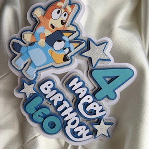 May include: A layered cake topper featuring the cartoon characters Bluey and Bingo, the number 4, and the text "HAPPY BIRTHDAY LEO". The design incorporates silver glitter accents, light blue, and dark blue cardstock layers with a white base.