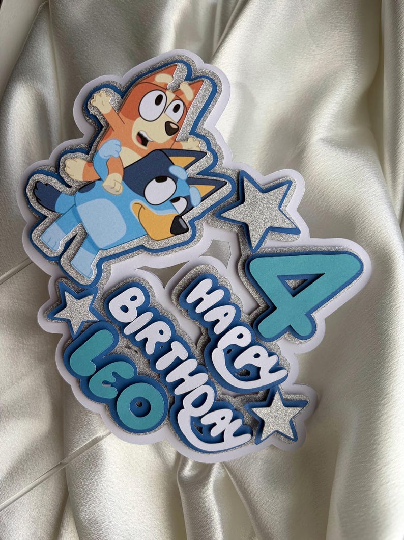 May include: A layered cake topper featuring the cartoon characters Bluey and Bingo, the number 4, and the text "HAPPY BIRTHDAY LEO". The design incorporates silver glitter accents, light blue, and dark blue cardstock layers with a white base.