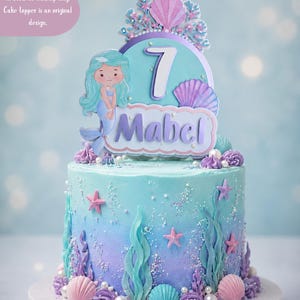 May include: A cake topper featuring a mermaid illustration, a large number 7, and the name Mabel in purple lettering. The design includes pink and purple seashells, coral accents, and white pearls, all set against a light blue background.