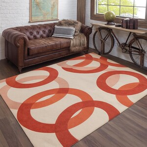 May include: A rectangular rug featuring a cream-coloured background and large overlapping circles in shades of orange and coral. The rug is on a dark wood floor in a living room, with a brown leather sofa in view.