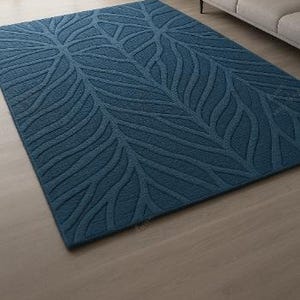 May include: A large, rectangular, teal-colored area rug with a leaf-like pattern. The rug is made of a soft material and is placed on a wooden floor. The rug is in a living room setting, with a sofa and window visible in the background.