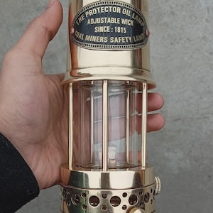 Op de afbeelding: Een messing Protector olielamp met verstelbare lont, ontworpen als kolenmijnveiligheidslamp sinds 1815. De lamp heeft een heldere glazen cilinder en wordt door een persoon vastgehouden. Een vintage item.