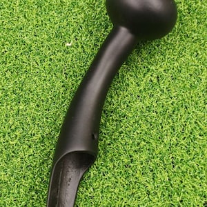 Peut inclure: Un objet métallique noir incurvé avec un dessus arrondi et une extrémité pointue. L'objet a une finition lisse et mate et semble être une poignée ou un outil. Il est posé sur un fond d'herbe verte artificielle.