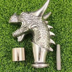 May include: A silver dragon head sculpture with detailed scales and horns. It is shown with a metal ring and a threaded rod. The items are displayed on a green surface. The dragon head is a decorative item, possibly for a door handle.