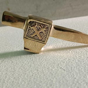 May include: A gold-coloured pickaxe-shaped ring. The ring has a rectangular head with an embossed design and a pointed blade extending from the side. The ring is set against a white surface.