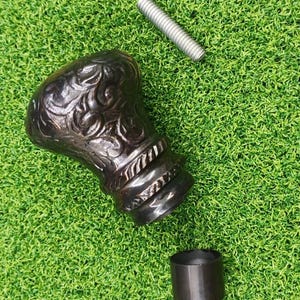 Puede incluir: Un mango de bastón ornamentado, de color marrón oscuro, con un diseño floral, una varilla roscada de metal y una férula de metal negro, todo sobre una superficie verde. El mango presenta un patrón tallado detallado y un acento en forma de cuerda.