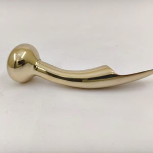 May include: A gold-colored, curved metal object with a rounded end and a pointed, flattened tip. The object has a smooth, reflective surface and appears to be a decorative or functional item, possibly a handle or a unique design element.