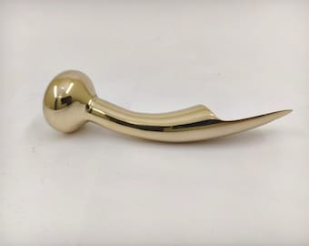 Puede incluir: Un objeto metálico dorado y curvado, con un extremo redondeado y una punta aplanada y puntiaguda. El objeto tiene una superficie lisa y reflectante, y parece ser un elemento decorativo o funcional, posiblemente un asa o un elemento de diseño único.