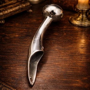 May include: A polished silver metal object with a rounded top and curved handle. The object is lying on a dark wooden surface. A lit candle in a gold candlestick and antique books are in the background.