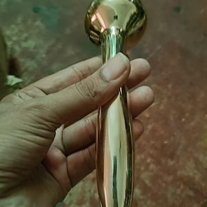 May include: A gold massage tool with a rounded head and a long, slender handle. The tool is held in a hand. The background is a muted green and brown.
