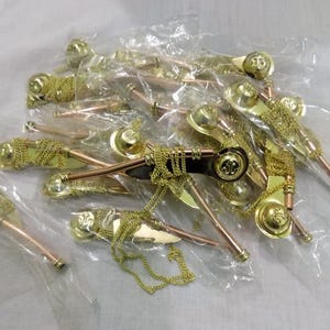 Puede incluir: Un conjunto de silbatos dorados con tubos de cobre y cadenas, empaquetados en plástico transparente. Los silbatos tienen un diseño decorativo y están dispuestos sobre una superficie blanca.