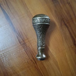 Puede incluir: Un sello de metal antiguo con un diseño floral ornamentado. El sello tiene una parte superior redondeada y un mango detallado y texturizado. El metal tiene un aspecto deslustrado y envejecido, lo que sugiere que es vintage. El sello probablemente se utiliza para sellar o marcar.