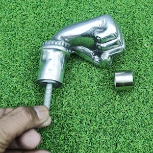 Puede incluir: Un tirador de puerta plateado con forma de puño cerrado. El tirador tiene un poste roscado y una pieza cilíndrica separada. El tirador se muestra sobre un fondo verde texturizado.