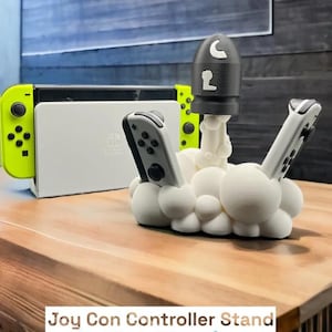 Peut inclure: Une console Nintendo Switch blanche et des manettes Joy-Con vert néon sont posées sur une surface en bois. Un support de manette blanc en forme de nuages maintient deux manettes Joy-Con. Une fusée noire est posée sur le support. Le texte en bas indique "Joy Con Controller Stand."
