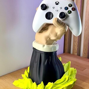 Peut inclure: Une manette Xbox blanche tenue par une main sculptée, sur une base noire avec un col crème et des accents verts vifs en forme de feuilles. La manette comporte des joysticks noirs et des boutons colorés.