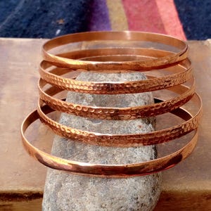 May include: A stack of five copper bangles, with three featuring a textured hammered finish and two having a smooth, polished surface. The bangles are arranged vertically, showcasing the metallic reddish-brown color and circular shape.