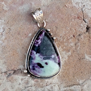 May include: A teardrop-shaped pendant with a silver-colored frame. The stone within the frame features a blend of purple, black, and pale green hues. The pendant has a decorative bail and small silver accents. The jewelry is suitable for necklaces.