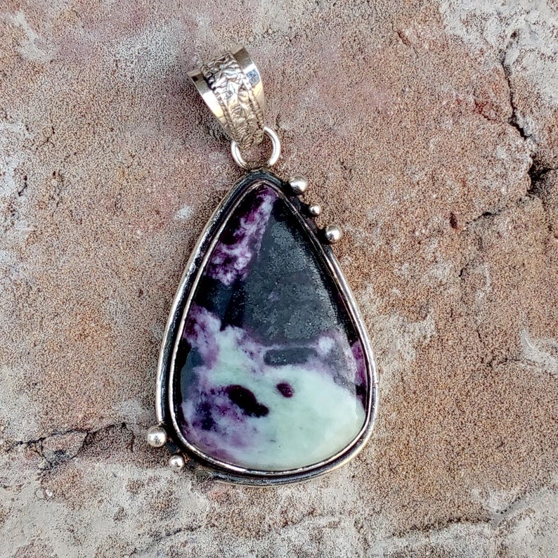 May include: A teardrop-shaped pendant with a silver-colored frame. The stone within the frame features a blend of purple, black, and pale green hues. The pendant has a decorative bail and small silver accents. The jewelry is suitable for necklaces.