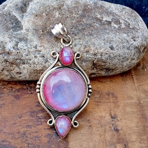 May include: A silver pendant with a large, round, pink and blue iridescent gemstone at the center. Two smaller pink gemstones are set above and below the central stone. The pendant has an ornate, curved silver frame.