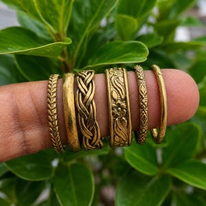 May include: A set of six gold-toned metal rings featuring various textures and patterns, including a chevron design, a smooth band, a braided rope pattern, a floral embossed band, a geometric diamond-shaped band, and a plain thin band.