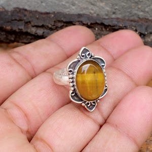Puede incluir: Anillo de plata con una piedra ojo de tigre ovalada, de color marrón dorado. La piedra está engastada en un engaste de plata decorativo y ornamentado. El anillo se muestra en una mano, mostrando el diseño y el color de la joya.