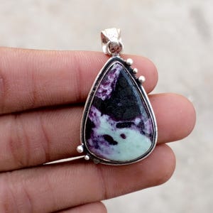 May include: A teardrop-shaped pendant with a silver-colored frame. The pendant features a polished stone with a mix of black, purple, and pale green colors. The pendant has a small loop at the top for a chain and decorative silver accents.
