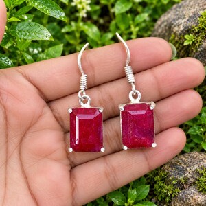 May include: A pair of silver drop earrings featuring rectangular, faceted deep red ruby gemstones. Each earring is suspended from a simple silver hook wire, with the stone held in a four-prong setting that secures the vibrant red gemstone in place.