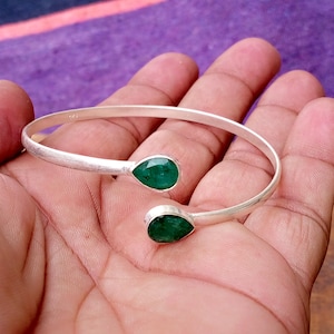 May include: A silver-colored cuff bracelet with two pear-shaped, vibrant green gemstones. The bracelet is open-ended and rests on a hand, showcasing the jewelry's design and color contrast.