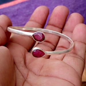 May include: A silver cuff bracelet with two teardrop-shaped, deep red gemstones. The bracelet has a simple, elegant design, with the gemstones set at each end of the open cuff. The gemstones are a rich, vibrant colour.