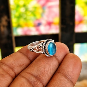 May include: A silver ring featuring an oval-shaped, translucent blue cabochon stone set in a twisted rope-style bezel. The band displays decorative scrollwork and open metal loops on the sides, with a polished finish throughout the metal structure.