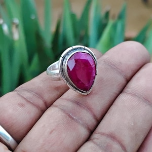 May include: A silver ring featuring a large, pear-shaped, faceted deep pink gemstone set in a smooth metal bezel. The band is plain and polished, showcasing a simple design that highlights the vibrant color and geometric cut of the central stone.