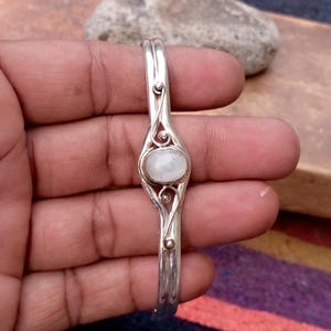 May include: A silver cuff bracelet featuring a central oval-shaped white moonstone cabochon. The metal band is constructed from two parallel silver wires that curve around the stone, accented with small silver spheres at the junction points.