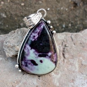 May include: A teardrop-shaped pendant with a silver-colored metal frame. The stone within the frame features a mix of black, purple, and pale green colors. The pendant has a decorative bail and small silver-colored accents.