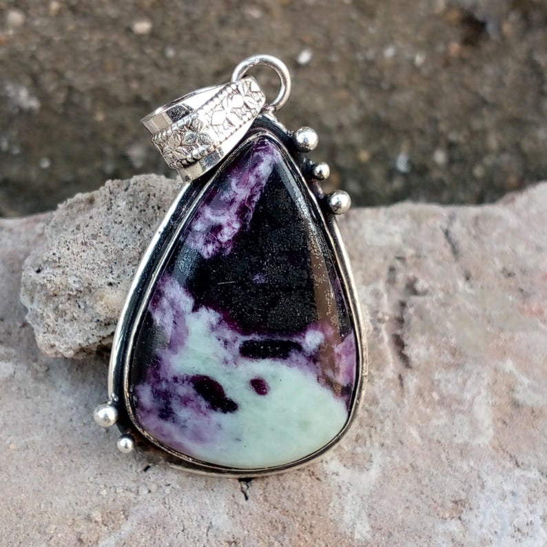 May include: A teardrop-shaped pendant with a silver-colored metal frame. The stone within the frame features a mix of black, purple, and pale green colors. The pendant has a decorative bail and small silver-colored accents.
