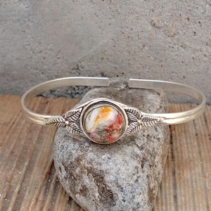 May include: A silver-toned cuff bracelet with a central, round stone featuring orange, red, and white hues. The stone is set in a decorative bezel with leaf accents. The bracelet is displayed on a gray rock against a wooden surface.