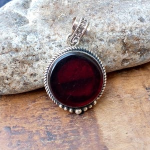 Könnte beinhalten: Runder Anhänger mit einem tiefroten Stein, der in einen silberfarbenen Metallrahmen gefasst ist. Der Rahmen hat ein gedrehtes Seildesign und kleine Perlenakzente. Die Öse ist verziert.