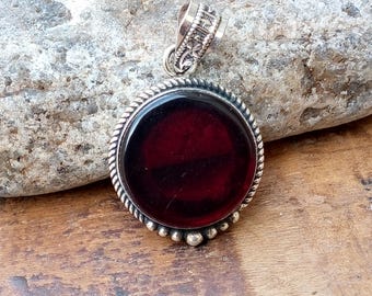 May include: A round pendant with a deep red stone set in a silver-colored metal frame. The frame has a twisted rope design around the stone and small beaded accents. The bail is ornate.
