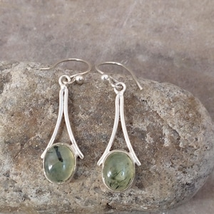 May include: A pair of silver earrings with oval, light green gemstones. The earrings have a teardrop design with a hook closure. The gemstones have dark green inclusions.