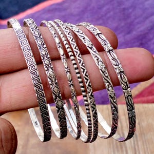 May include: A collection of silver bangles with intricate patterns. The bangles feature various designs, including floral, geometric, and wave-like motifs. The silver jewelry is displayed against a purple and brown background.
