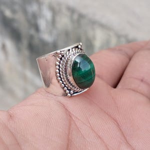 Puede incluir: Anillo de plata con una gran piedra ovalada de malaquita pulida. El anillo tiene una banda ancha con detalles decorativos de cuentas y cuerda alrededor del engaste de la piedra. La malaquita es de color verde oscuro con vetas visibles.