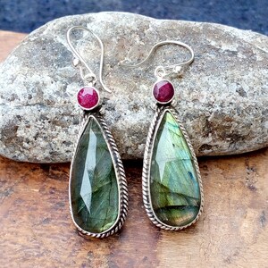 May include: A pair of silver earrings with teardrop-shaped labradorite stones. Each earring features a small, round, red gemstone at the top and a braided silver border around the labradorite. The labradorite stones display green and blue iridescence.