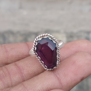 Puede incluir: Un anillo de plata con una gran piedra preciosa facetada de color púrpura oscuro. La piedra preciosa está engastada en un borde decorativo retorcido en forma de cuerda. El anillo se sostiene en una mano, mostrando el diseño y el color.