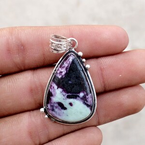 May include: A teardrop-shaped pendant with a silver-colored frame. The pendant features a stone with a mix of black, purple, and pale green colors. The top of the pendant has a decorative bail for a chain. Small silver beads accent the frame.