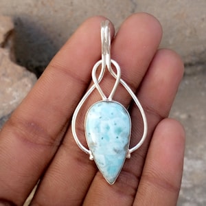 May include: A teardrop-shaped pendant with a light blue and white stone, set in a silver metal frame. The stone has a unique pattern of blue and white swirls. The pendant is suspended from a silver bail.