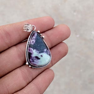 May include: A teardrop-shaped pendant with a silver bail and setting. The stone features a mix of dark purple, black, and light green colors. Small silver beads adorn the top of the pendant.