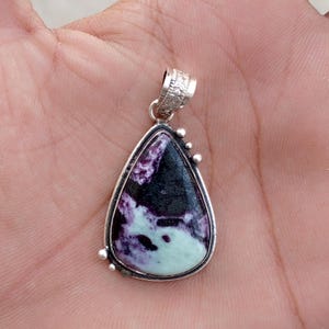 May include: A teardrop-shaped pendant with a silver-colored frame. The pendant features a polished stone with a mix of black, purple, and pale green colors. The bail is decorated with a floral pattern. Small silver beads adorn the frame.