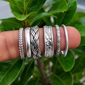 May include: A set of six silver-toned rings featuring various designs including a chevron pattern, a smooth band, a braided texture, a floral motif, a geometric diamond pattern, and a plain thin band, all displayed in a row on a finger.