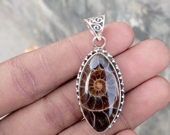 May include: Oval-shaped pendant featuring a polished, fossilized ammonite encased in a silver-toned setting. The pendant showcases the intricate spiral pattern of the fossil, with a decorative bail at the top. The ammonite is a rich brown color.